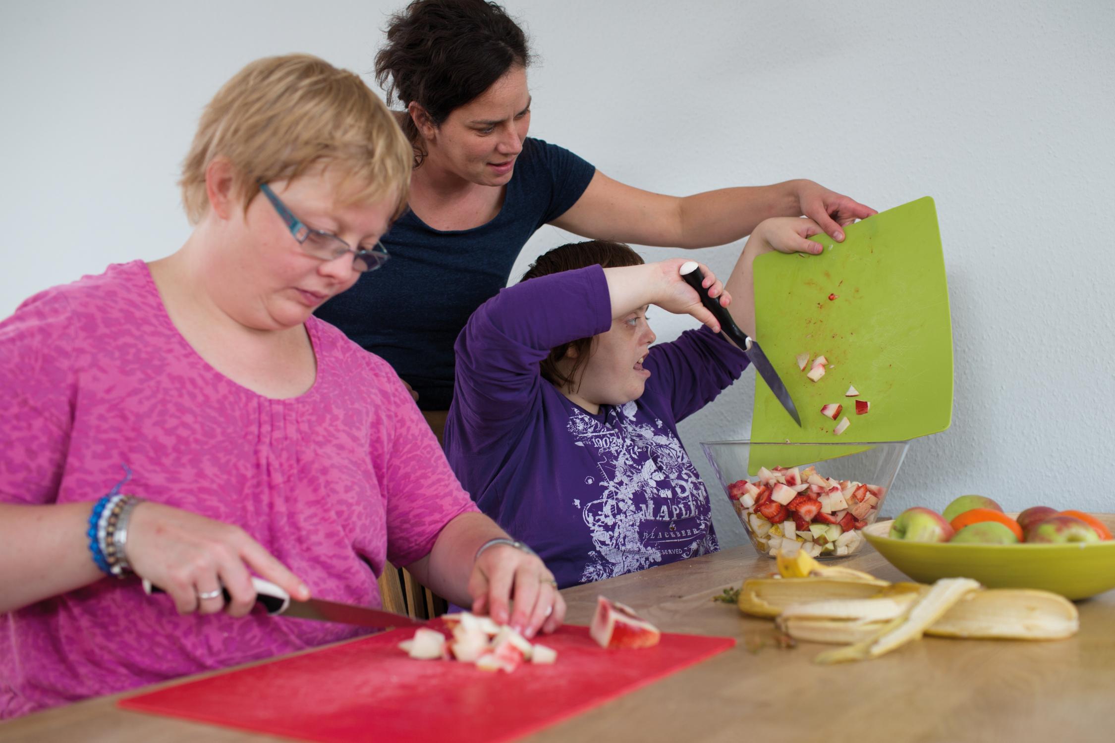Eine Frau schneidet Obst, während eine andere Frau das geschnittene Obst mit Hilfe einer dritten Person in eine Schüssel schüttet.