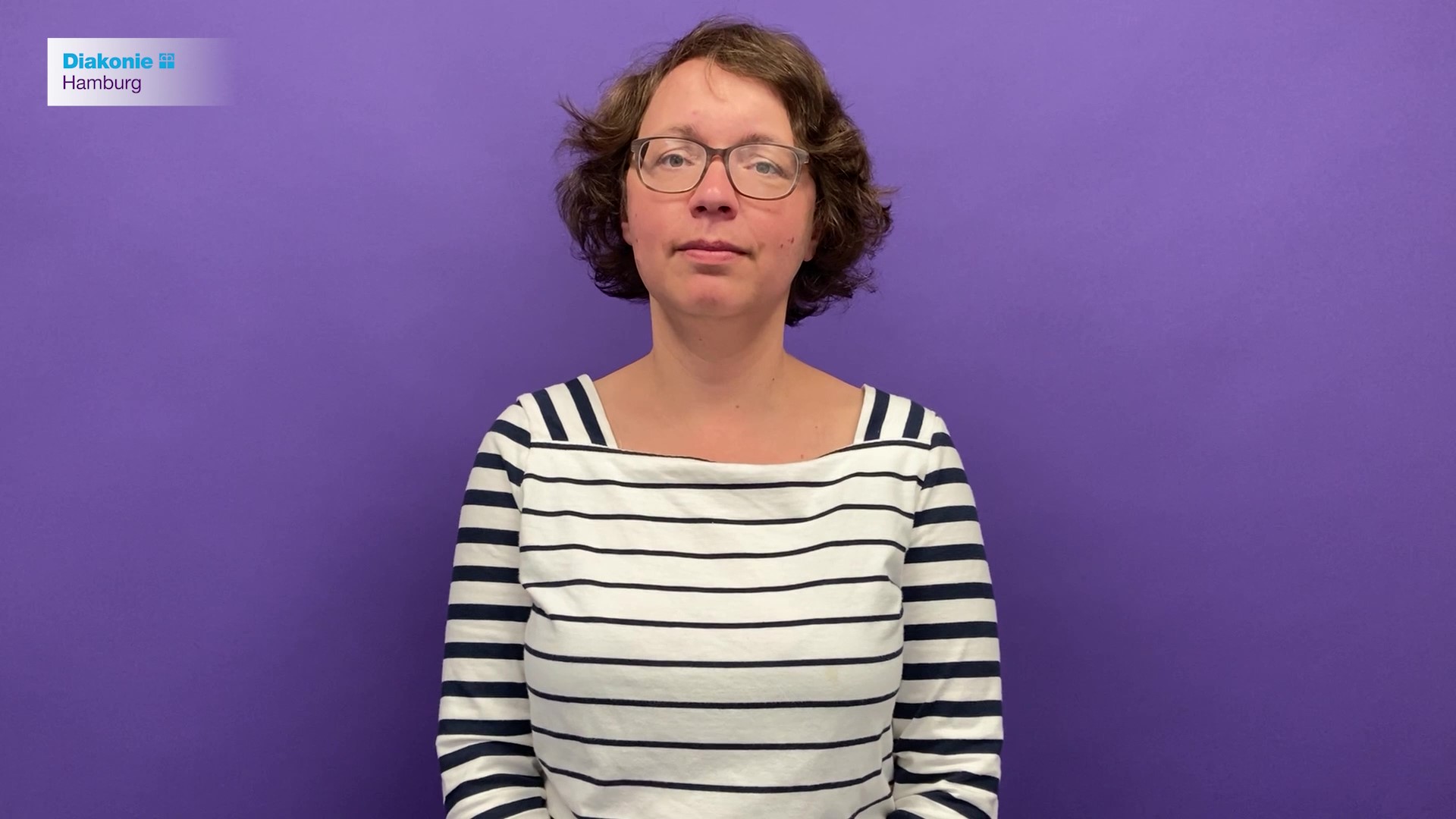 Eine Frau mit kurzen braunen Haaren und Brille steht vor einem lila Hintergrund.